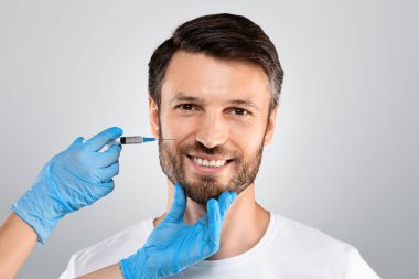 Kozmetik uzmanları koruyucu eldivenlerle orta yaşlı bir adamın yüzünü dolduruyor. Gri stüdyo arka planı, fotokopi odası. Gülümseyen adam güzellik salonunda ya da klinikte yaşlanmayı önleme prosedürü uyguluyor.