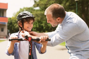 Güvenlik kavramı. Gülümseyen yetişkin babanın portresi parkta küçük çocuğa koruma kaskı takıyor cesur oğluna elektrikli scooter sürmeyi öğretiyor. Hafta sonu etkinliğimiz