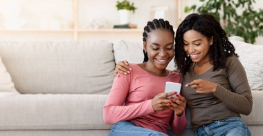 Mutlu siyahi bayanlar evde dinlenirken yeni mobil uygulama kullanıyorlar, boş yer. İki neşeli Afro-Amerikalı kız kardeş akıllı telefondan sosyal medyayı kontrol ediyor, oturma odasında yerde oturuyor.
