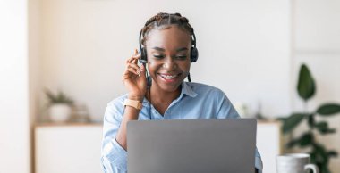 Neşeli siyah kadın yönetici kulaklık takıyor ve ofiste dizüstü bilgisayar kullanıyor, müşteriye danışıyor, müşteriyle konuşuyor, boş alan yaratıyor.