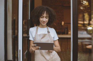 Covid-19 karantinası sırasında yeni normal, sosyal mesafeler ve çevrimiçi düzen. Önlüklü Afrikalı Amerikalı genç bir kadın internette sörf yapmak için tablet kullanıyor ve müşterileri giriş kapısının yanında bekliyor.