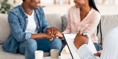 Evlilik danışmanı mutlu siyah çiftle başarılı terapi seanslarından sonra notlar alıyor. Genç eşler kanepede el ele oturuyorlar. Uzlaşma sonrası el ele tutuşuyorlar.