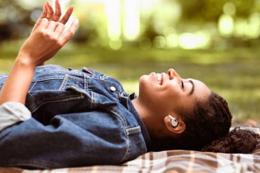 Müzik aşığı. Gevşemiş Afro-Amerikan Kadın Kulaklıkla Favori Şarkısını Dinliyor Ekose Üzerinde Uzanıp Dışarıda Parkta Hafta Sonu Keyifli Geçiriyor. Rahatlama Boş Konsepti