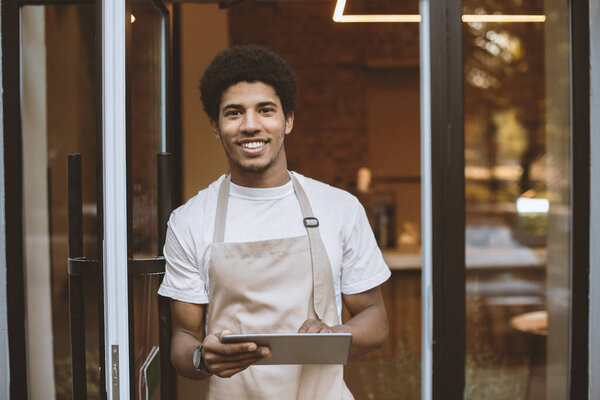 Современный малый бизнес, кафе и работа на свежем воздухе, новая нормальная и современная. Портрет счастливого африканского мужчины тысячелетия в фартуке с табличкой стоит у входной двери в ресторане на вынос утром