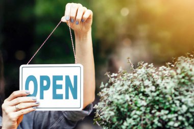 Opening small business after covid-19 pandemic and positive human emotions. Smiling young beautiful lady in apron turns sign with inscription is open, on glass door in flower studio with plants