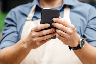 Satış bitkileri, dekor, hediyeler ve başlangıç. Önlüklü ve koruyucu maskeli meşgul bir adam mesaj yazıyor ya da sosyal ağlarda internette sörf yapıyor çiçekçi dükkanının yanındaki akıllı telefondan, kapatılıyor, kırpılıyor.