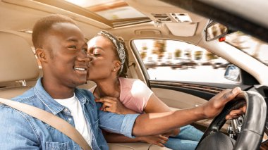 Arabayla seyahat ederken gülümseyen kocasını öpen siyahi kadını sevmek, kopyalamak. Mutlu Afro-Amerikan aşıkları hafta sonu birlikte seyahatin tadını çıkarıyorlar, arabayla tatile çıkıyorlar.