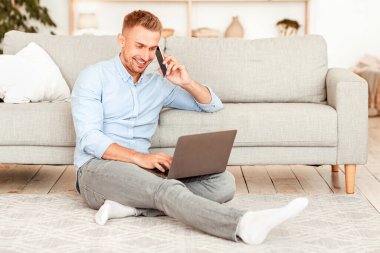 Ev İşyeri. Oturma odasında çalışan genç serbest çalışan, yerde dizüstü bilgisayarla oturan, akıllı telefondan konuşan. Kurumsal İletişim Konsepti. Neşeli adam telefon ediyor, bilgisayar kullanıyor.