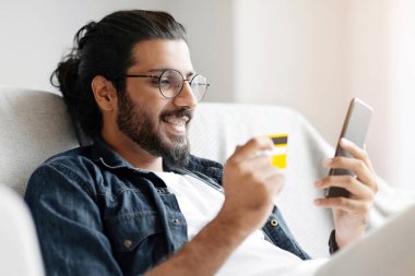 Evinde akıllı telefon ve kredi kartıyla alışveriş yapan neşeli, yakışıklı bir Arap adam, gözlüklü ve diş telli Milenyum adamı, İnternet 'te döşeme keyfi yapıyor, oturma odasında koltukta oturuyor.