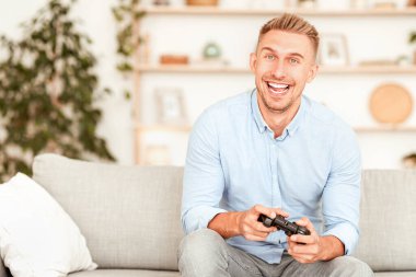 Mens Hobi Konsepti. İnternette oynayan gülümseyen yetişkin oyuncunun portresi, joystick oyun alanı kullanıyor. Duygusal adam video oyunlarından hoşlanıyor, oturma odasındaki kanepede oturuyor, monitöre bakıyor.