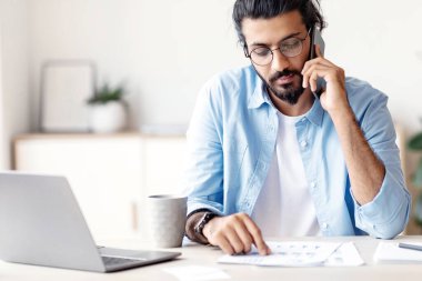 Genç Arap erkek girişimci cep telefonuyla konuşuyor ve modern ofiste kağıtlarla çalışıyor, masamızda oturan yakışıklı bir doğulu adam mali raporları kontrol ediyor ve cep telefonuyla konuşuyor, boş yer var.