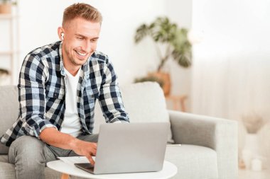 Eğitim Konsepti. İnternette çalışan neşeli adam dizüstü bilgisayarla koltukta oturuyor ve kulaklık takıyor.