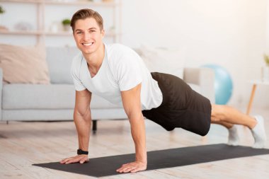 Formda kal ve evde egzersiz yap. Gülümseyen kaslı adam oturma odasının içinde şınav çekiyor.