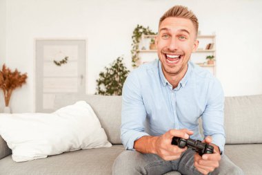 Bekarlar için eğlenceli bir hobi. İnternette video oyunu oynayan, elinde joystick tutan çok mutlu bir adam. Oturma odasındaki kanepede oturan erkek oyuncunun portresi. Kapalı, bulanık arka plan.