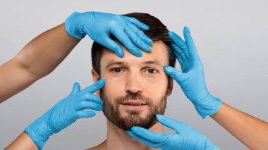 Plastik cerrah koruyucu eldivenleriyle gri stüdyo geçmişinde orta yaşlı bir adamın yüzüne dokunuyor. Estetik cerrahi ve estetik kozmetik konsepti. Çekici adam estetik ameliyata hazır.