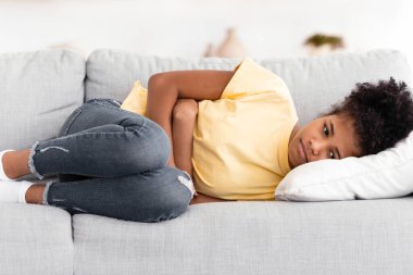 Siyahi bir genç kız midesi ağrıyan periyotlardan muzdarip. Kapalı koltukta karnına dokunarak yatıyor. Karın Ağrısı Konsepti