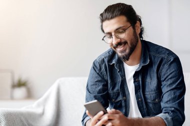 Yakışıklı Hintli genç adam elinde akıllı telefonuyla evde oturuyor, arkadaşlarıyla mesajlaşıyor, koltukta cep telefonuyla dinleniyor, yeni uygulamaları kontrol ediyor, mobil iletişimden keyif alıyor ya da sosyal ağlara takılıyor.