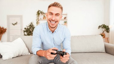 Ev Kavramında zaman geçirmek. Online video oyunları oynayan, elinde joystick tutan mutlu bir adamın portresi. Oturma odasındaki koltukta oturan yetişkin oyuncu, kameraya bakıyor, arka planı bulanık.