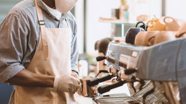 Covid-19 karantina, yeni müşteri hizmetleri kuralları ve sosyal mesafeler. Koruyucu maskeli ve eldivenli genç Afrikalı Amerikalı kahve makinesinde latte yapıyor.