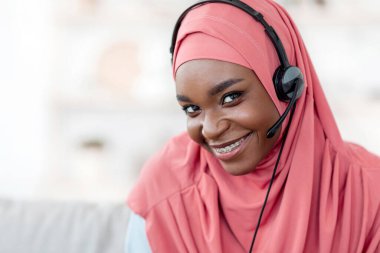 Müslümanlar için uzak görevler. Türbanlı Siyahi Kadın 'ın yakın plan portresi ve evdeki koltukta oturan kulaklıklı Afrikalı çağrı merkezi operatörü gülümseyen ve kameraya bakan kadın,