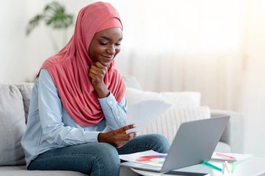 Sırıtkan Müslüman kadın tesettürde dizüstü bilgisayar ve renk paleti kullanıyor, yeni dairesi için iç tasarım seçiyor, dalgın Afrikalı İslamcı Kadın Tutma Örnekleri alıyor, Evdeki koltukta oturuyor,