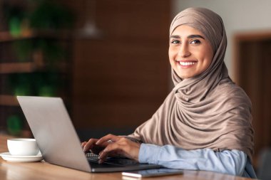 Kafe 'de çalışan tesettürlü neşeli Müslüman kadın dizüstü bilgisayar kullanıyor, kahve veya çay içiyor, fotokopi çekiyor. Yan bakışta gülümseyen Arap kadın dizüstü bilgisayarda yazı yazıyor, uzak işinden zevk alıyor.