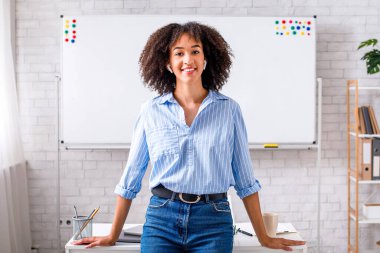 Öğretmen ve İngilizce dersleri evden çevrimiçi. Dost canlısı Afro-Amerikan kız, karatahtanın önünde, iç mekandaki kameraya bakıyor.