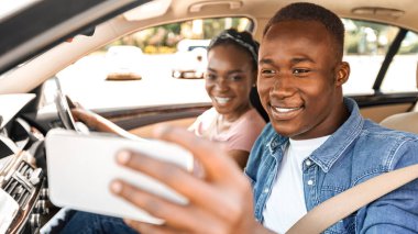 Mutlu genç siyah çift araba kullanıyor ve telefondan selfie çekiyor. Neşeli Afro-Amerikan milenyum erkeği ve kadın cep telefonuyla fotoğraf çektirirken rahat bir otomobille tatile çıkıyor, uzayı kopyalıyor.