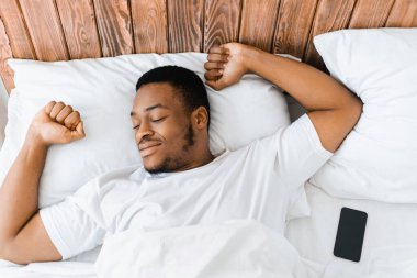 Dinlenmiş Afrikalı adam, uykusundan uyanıyor, yatağında uzanıyor, sabahları sıcak yatak odasında ellerini uzatıyor, gözleri kapalı gülümsüyor. İyi Dinlenme, Uyku ve Dinlenme Konsepti. Üst Görünüm