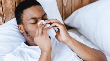 Evdeki yatakta yatan Rhinitis 'li hasta siyah adam kağıt mendilde akan burnunu siliyor. Sinüzit, grip ve soğuk algınlığı belirtileri gösteren hapşıran hasta Afrikalı adam. Panorama