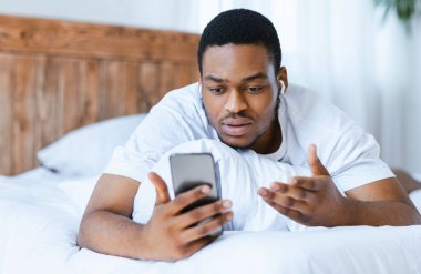 Ciddi Afro-Amerikan bir adam akıllı telefonuyla video görüşmesi yapıyor. Evde yatakta yatıyor. Uzak Çevrimiçi İletişim, Video Konferansı Mobil Uygulama Konsepti. Seçici Odaklanma