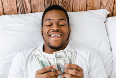Mutlu Afrikalı Amerikalı adam para sayıyor nakit para sayıyor evdeki yatak odasında yatıyor. Finansal Zenginlik, Bütçe Geliri ve Mali Tasarruf Güvenliği. Seçici Odaklanma, Görünümün Üzerinde
