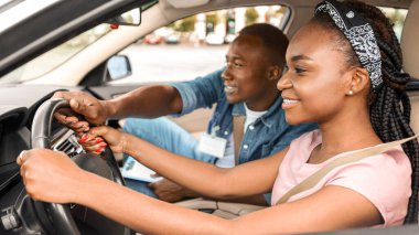 Gülümseyen genç siyahi kadın sürücü kursunda park pratiği yapıyor, yakışıklı bir eğitmenle arabada oturuyor. Neşeli Afro-Amerikalı adam, araba sürmede kız öğrenciye yardım ediyor.