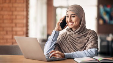 Müslüman bayan girişimci kafede dizüstü bilgisayarla çalışıyor, cep telefonuyla iş görüşmesi yapıyor, boşluğu kopyalıyor.