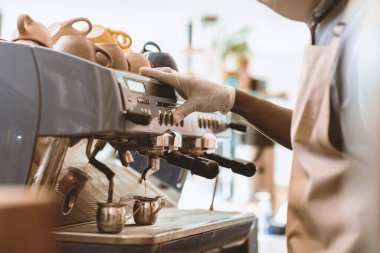 Kafeterya ve covid-19 salgınındaki modern kahve malzemeleri. Koruyucu maskeli, önlüklü ve eldivenli genç Afrikalı Amerikalı adam kafede espresso makinesiyle çalışıyor.