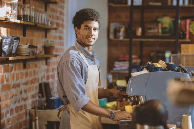 Modern kafeteryada baristalık, küçük işletme sahibi ve restoran açılışı. Gülümseyen bin yıllık Afro-Amerikan önlüklü adam kahvesini kahve tezgahında yapıyor.