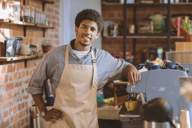 Sabah favori kafede dostça bir toplantı. Önlüklü, gülümseyen Afrikalı Amerikalı barmen barda espresso makinesinin yanında duruyor ve kafeteryanın içindeki boş alana bakıyor.