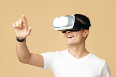 Adam sanal gerçeklik kulaklığıyla eğleniyor. Beyaz tişörtlü ve sanal gerçeklik gözlüklü neşeli genç çocuk kum arka planda izole edilmiş boş alanda sanal düğmeye basıyor.