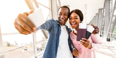 Maceraya hazırım. Havalimanı terminalinde Smartphone ile selfie çeken neşeli siyah bir çiftin portresi, kamerada birlikte poz vermeleri, pasaport ve biletleri ellerinde tutmaları,