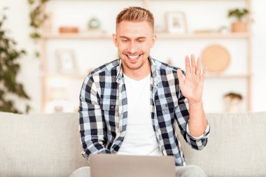Çevrimiçi Video Çağrısı. Bilgisayarda sanal konferans yapan gülümseyen adamın portresi, web kamerasına el sallıyor. İletişim Konsepti. Adam bilgisayarla sohbet ediyor, evdeki koltukta oturuyor.