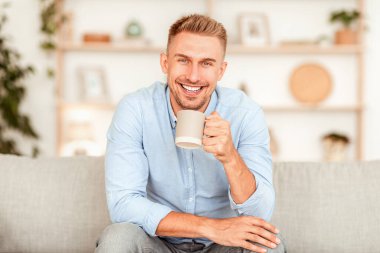 Erkenci. Gülümseyen adam evde kahve içiyor ve kameraya bakıyor, kanepeye oturuyor,