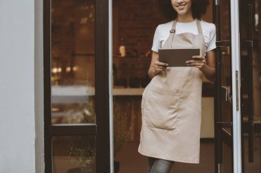 COVID-19 salgınından sonra çevrimiçi sipariş vermek için modern cihazlar, yeni normal ve müşteri hizmetleri. Afro-Amerikan önlüklü neşeli genç bayan tabletle sabah kafenin ön kapısında duruyor.