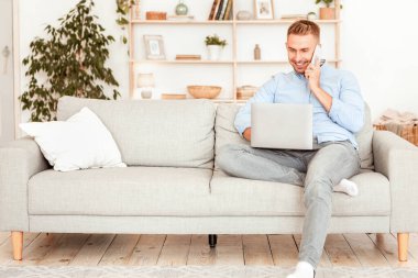 Kurumsal İletişim Konsepti. Neşeli adam telefonla konuşuyor, dizüstü bilgisayarda yazıyor, oturma odasında kanepede oturuyor. Telekomünikasyon ve telesekreter. İş için bilgisayar kullanan kendinden emin bir yetişkin.