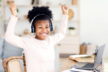 Bilgisayarda yumruklarını sallayan neşeli siyah kız çevrimiçi derste not alıyor evde uzaktan öğreniyor, kulaklık takıyor.