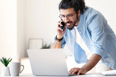 Gülümseyen Doğu 'lu genç girişimci cep telefonuyla konuşuyor ve modern ofiste dizüstü bilgisayarla çalışıyor, masanın yanında duruyor, yakışıklı bir Arap çalışanı bilgisayardaki bilgileri kontrol ediyor, boş bir alan.