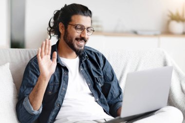 Çevrimiçi İletişim. Evde dizüstü bilgisayarla video görüşmesi yapan, web kamerasına el sallayan, genç bir Arap 'ın oturma odasında boş bir odada otururken birini selamlaması.