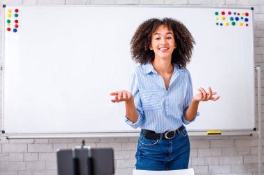 İş, öğrenci çevrimiçi ve kendini izole etme. Dost canlısı Afro-Amerikan milenyum bayanı oturma odasındaki takipçilere İngilizce öğrenme videosu kaydediyor. Beyaz tahta, dizüstü bilgisayar ve akıllı telefon ile.