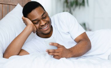 Neşeli siyahi adam hafta sonu sabahı evde yatarken akıllı telefon kullanıyor. Mobil Uygulama Kavramı