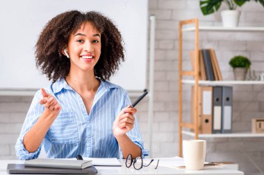 Karantina ve uzak dersler sırasında öğrenciler için İngilizce öğretmeni ya da öğretmeni. Dost canlısı Afro-Amerikan kadın gündelik dilin kurallarını açıklıyor. Masada dizüstü bilgisayarla oturup kameraya bakıyor.