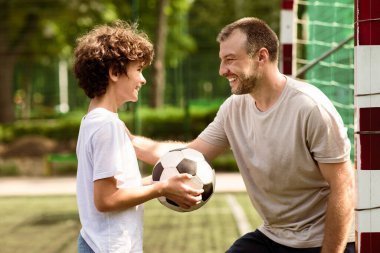 Çocuklar ve Açık Hava Aktivite Konsepti. Futbol topu tutan, gülümseyen kıvırcık çocuğun yan görüntüsü, omzuna hafifçe vurup okşayan, aile sahada futbol oynayan,